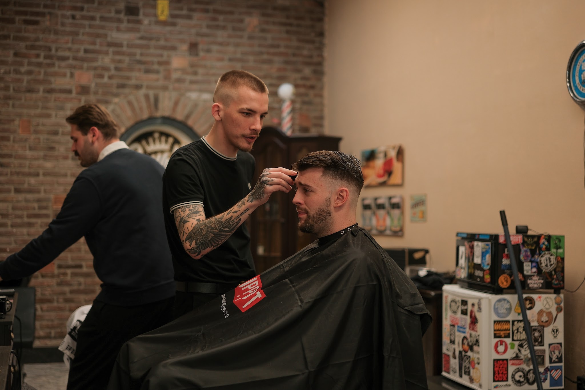 Barber werkt een fade bij voor de bakstenen muur, een tweede barber erachter